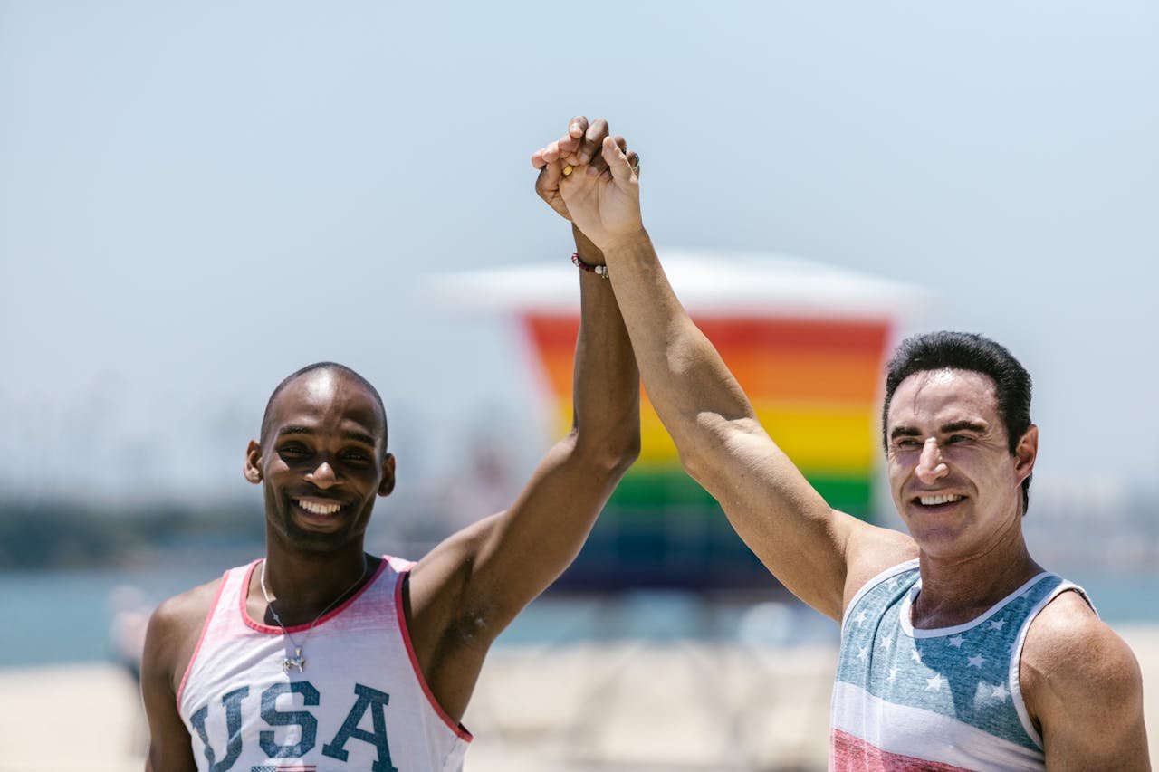 Home Interracial gay couple joyfully holding hands at a beach with rainbow colors in the background.