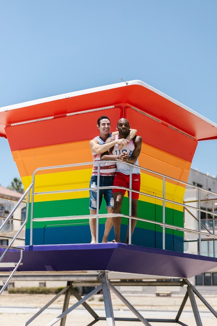 Happy interracial gay couple embracing on a colorful rainbow-painted lifeguard tower, celebrating love and diversity.