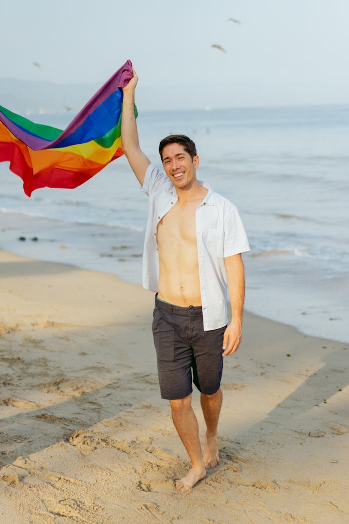 Cheerful man walking on the beach holding a rainbow flag, showing happiness and pride.